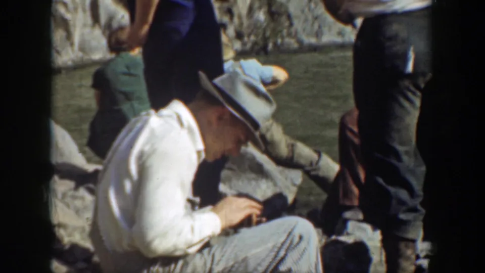 A man in vintage casual clothing sits near the rocky water's edge while visitors stand around him in the California outing scene.