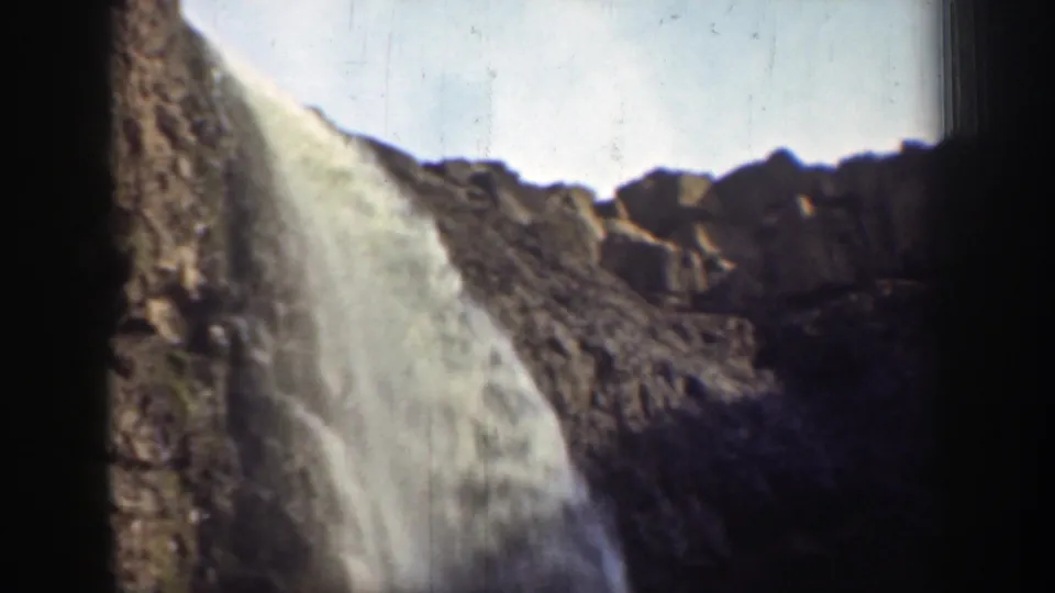 1937 California home-movie frame with water spilling over a cliff ledge against rugged rocks and a bright patch of sky.