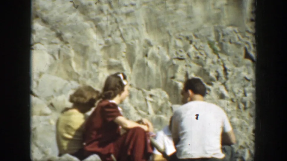 1937 California home-movie frame showing several tourists seated on rocks near a rugged stone wall.