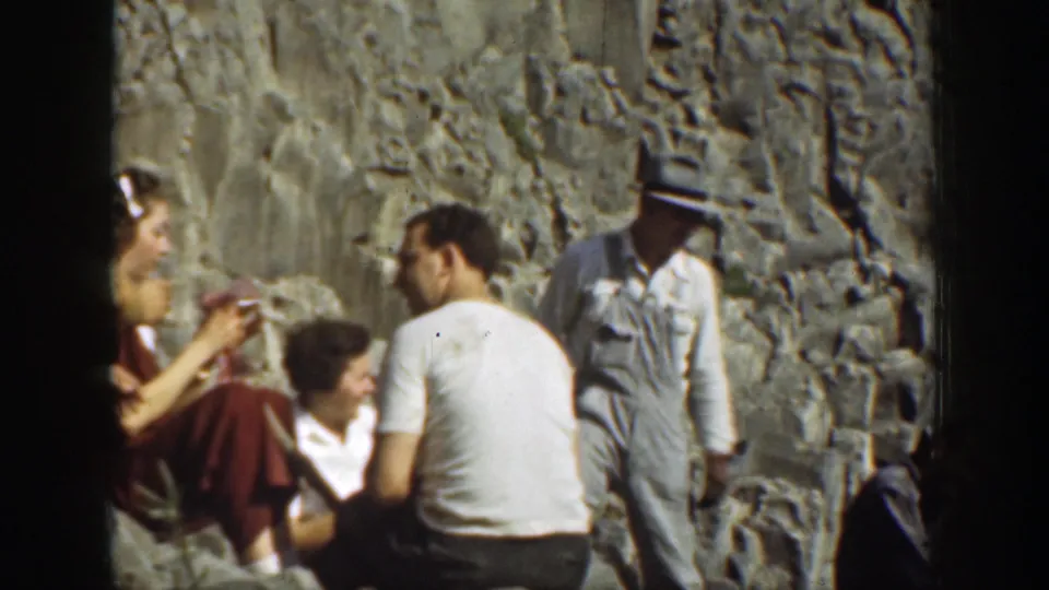 Tourists in period clothing sit and stand on rocks in 1937 California waterfall footage.