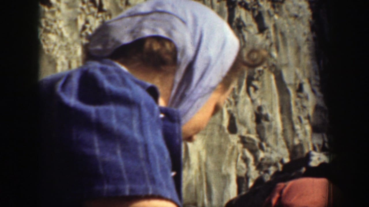 Woman in blue headscarf turned away beside a rocky cliff face.