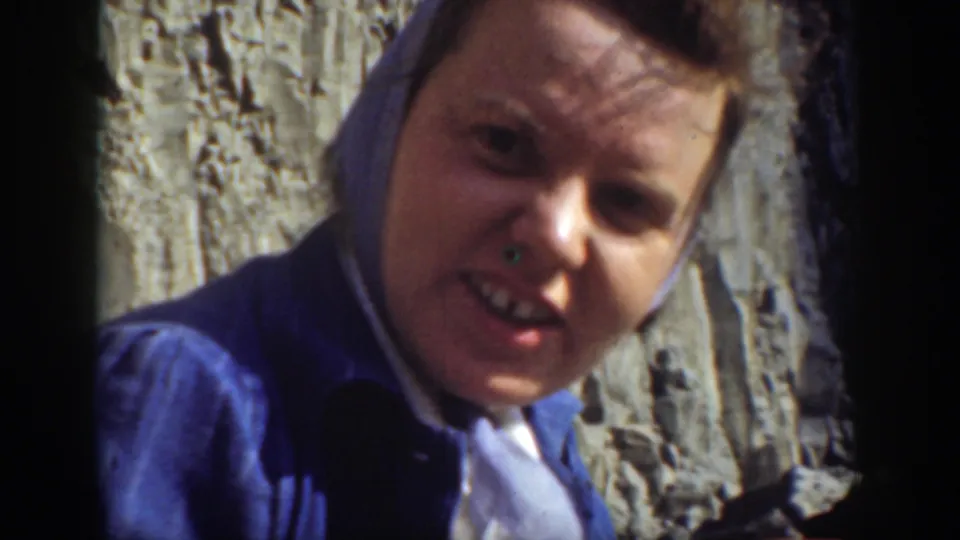 A woman in a blue headscarf smiles toward the camera near a sunlit rocky cliff in 1937 California home-movie footage.