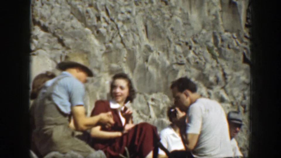 Adults in period casual clothing sit together near a California cliff face, including a man in a cap and a woman in a red dress.