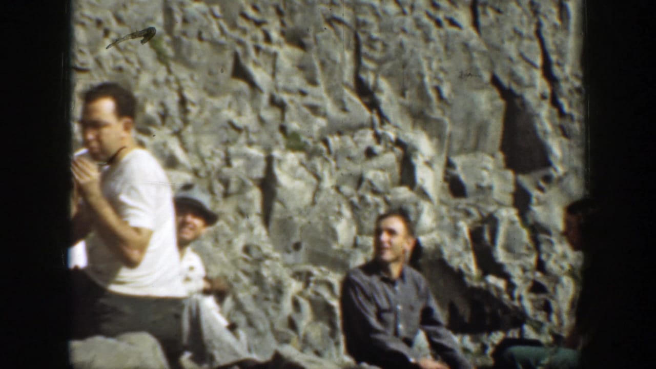 Men sit beside a rugged rock wall while one man in a white shirt smokes.