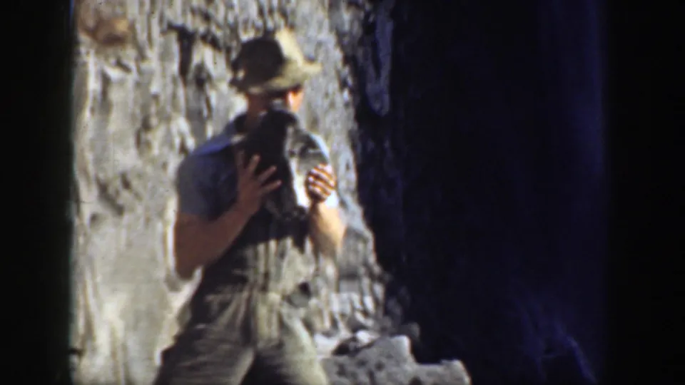 1937 California 8mm frame of a hat-wearing man holding a large stone beside a dark cave-like opening in a rocky setting.