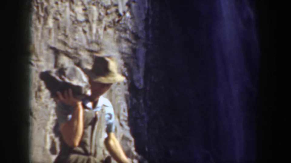 1937 California 8mm frame showing a man in a hat and vest raising a large stone beside a shadowed rocky opening.