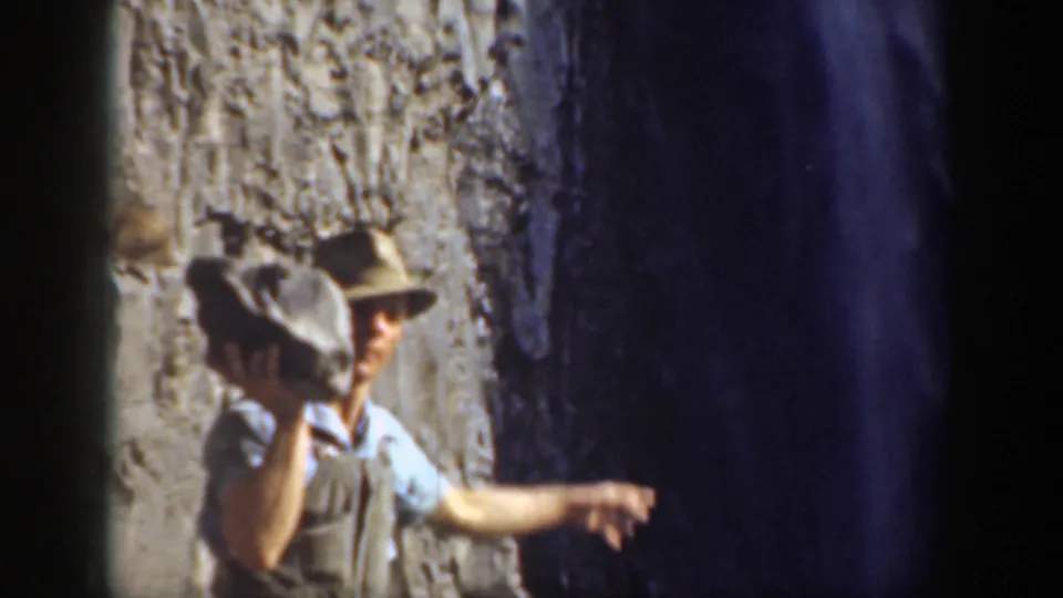 1937 California 8mm frame of a man holding a large stone while gesturing toward a dark cave-like opening in the rocks.