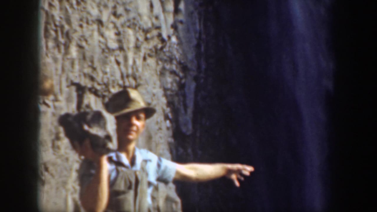 Man in a brimmed hat holds a rock beside a rugged stone wall.