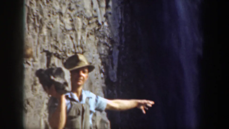 In 1937 California home-movie footage, a man in a light shirt and hat holds a rock near a steep rocky slope.