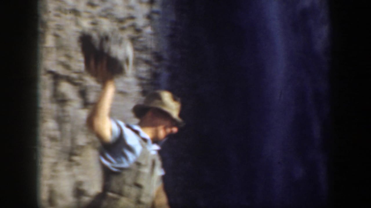 Man in a hat raises his arm beside a steep rock face.