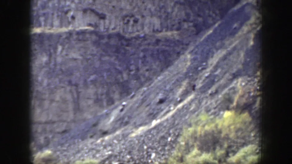 Long-distance 8mm view of a rugged California slope with layered rock, scrub, and small indistinct bright shapes on the hillside.