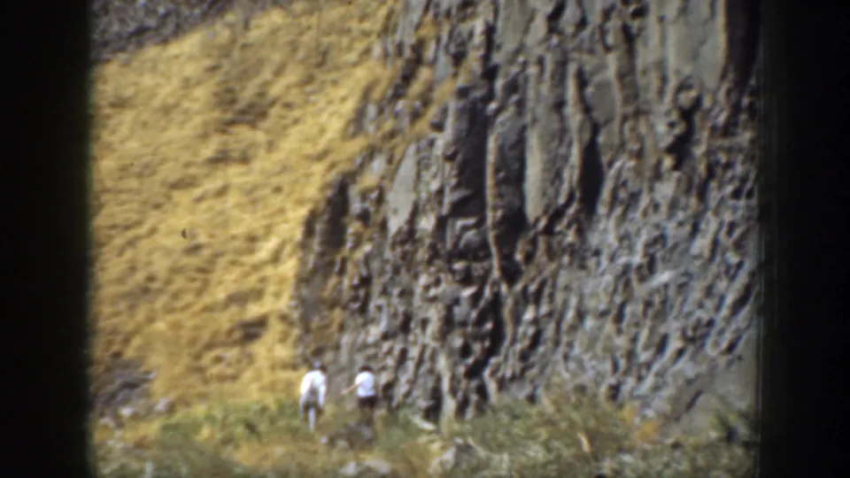 Two distant hikers in light clothing ascend a dry California hillside beside a dark layered rock face in 1937 home-movie footage.