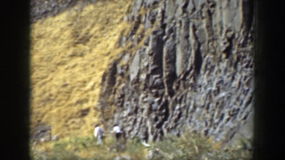 1937 California 8mm frame of two distant hikers moving along a dry hillside next to a textured layered rock wall.