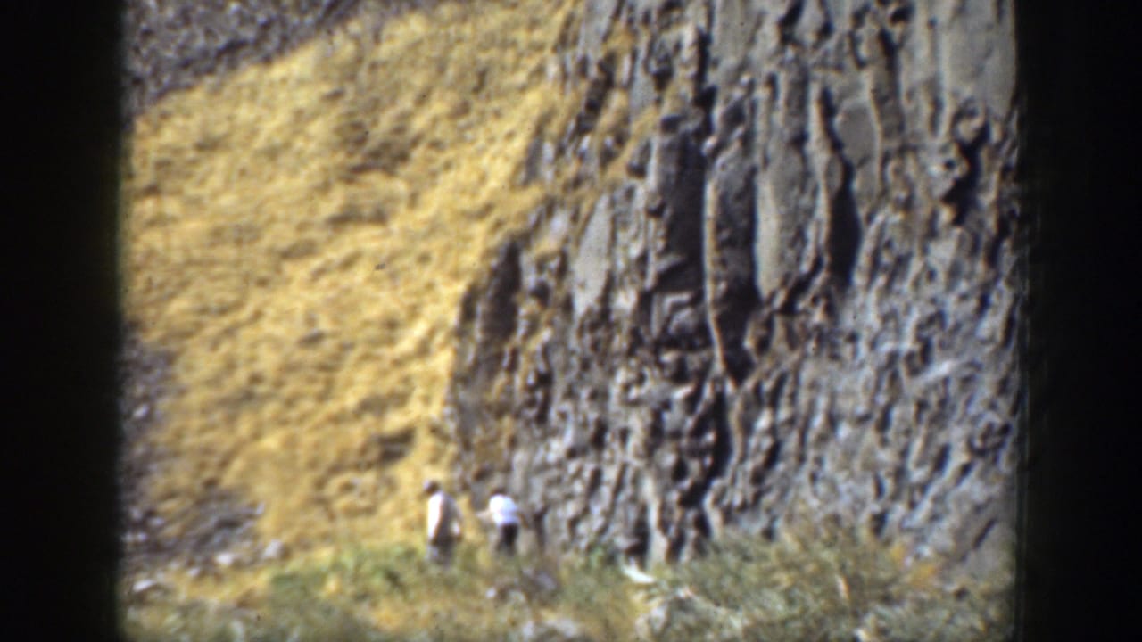 Two small walkers near a steep rocky cliff and dry grass slope.