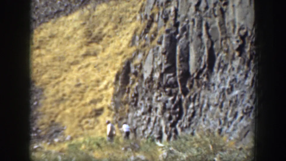 1937 California 8mm frame showing two distant people walking near a rocky cliff face with dry hillside grass.