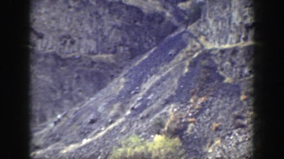 Soft 8mm view of a rugged California slope with visible trail lines, rock debris, and scattered green brush.