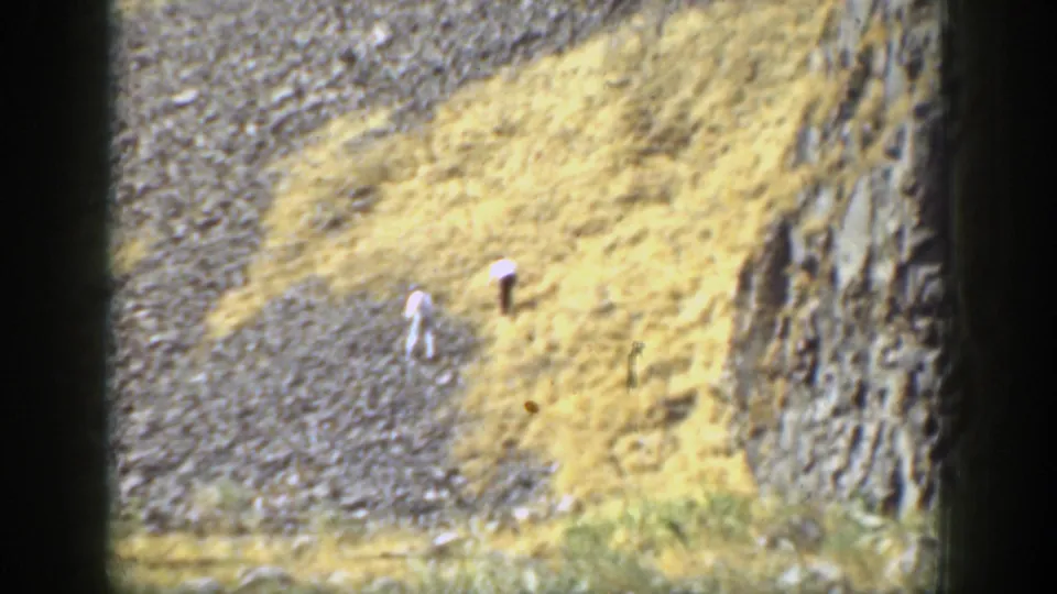 Two distant people appear to move across a steep rocky California slope, with dry grass and cliff-like rock visible nearby.