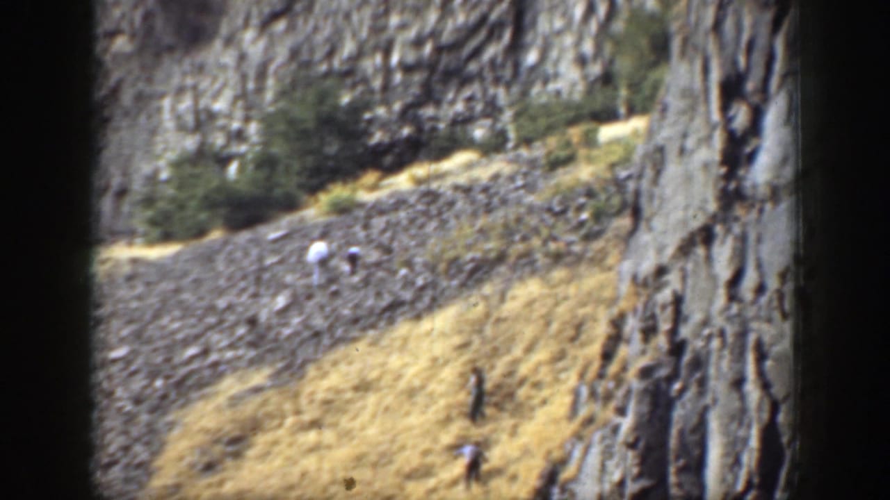 Distant hikers on a rocky slope below tall pale cliffs and sparse trees.