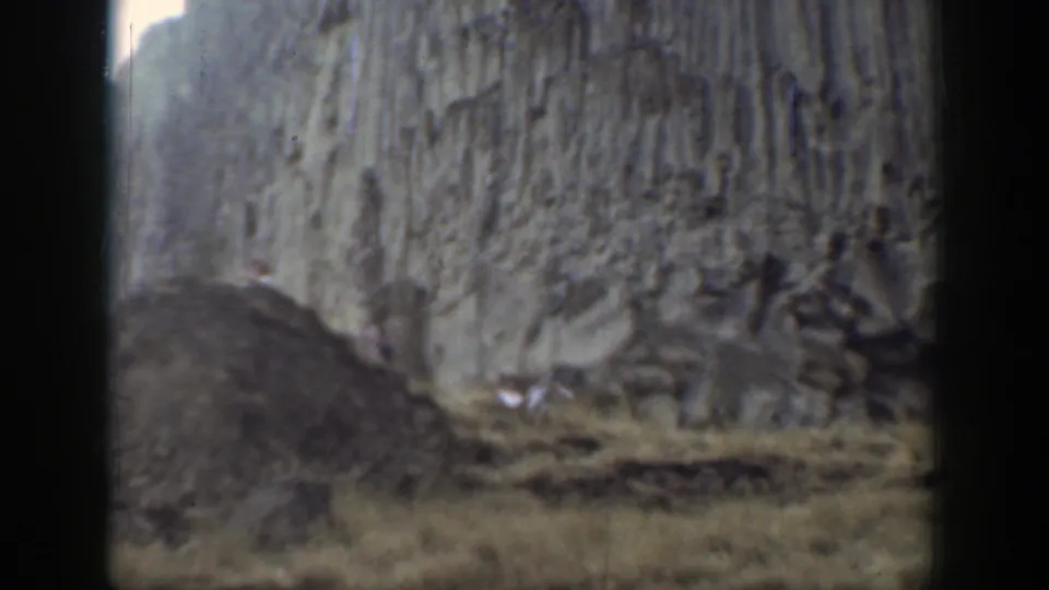 Soft 8mm frame of a rocky California slope, with a pale cliff face, loose stone mound, and dry grass foreground.
