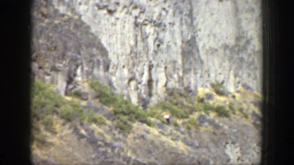 California 1937 home-movie frame with tiny hikers among shrubs and rocks below a high, weathered cliff face.