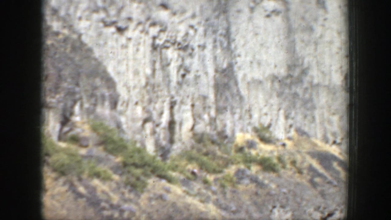 Steep pale cliff face above a rocky slope with sparse shrubs and dark film borders.