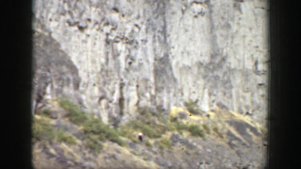 1937 California frame with a steep cliff, dry sloped terrain, low shrubs, and a tiny unidentified person near the rock face.