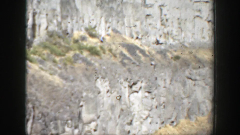 1937 California 8mm frame showing a striated rock face, scrubby slope, dry grasses, and visible film dust and borders.