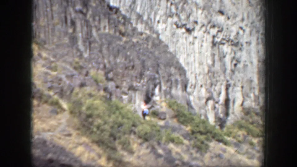 1937 California home-movie frame showing a distant walker in a white shirt on a narrow rocky trail below canyon formations.