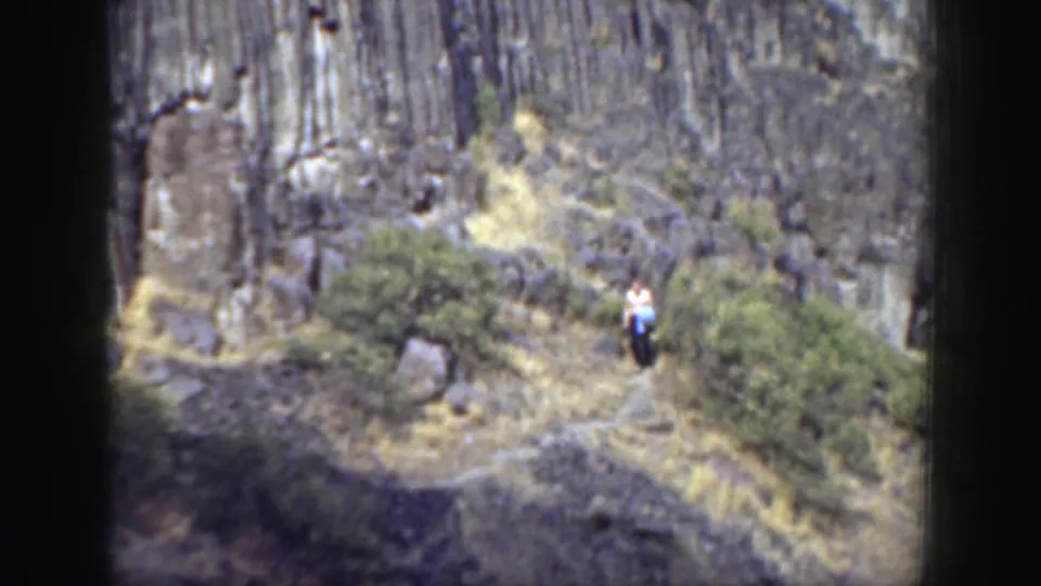 Distant figure in a white shirt appears on a rugged California canyon trail, surrounded by shrubs and layered grey rock.