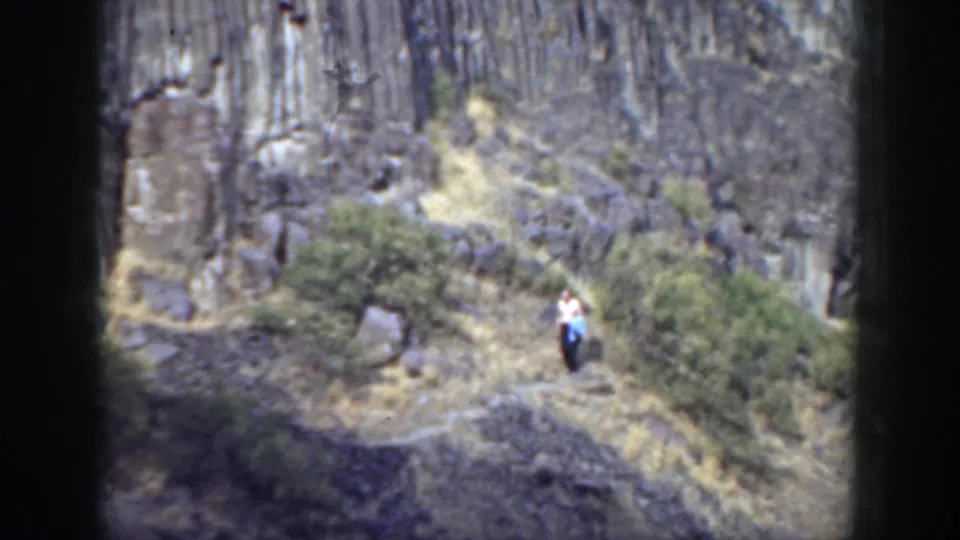 1937 California frame of a person in a white shirt and dark pants moving down a narrow trail through grey rocks and brush.