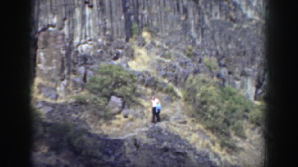 In 1937 California footage, a man in a light shirt stands on a narrow rocky trail with dry grass and basalt-like cliffs behind him.