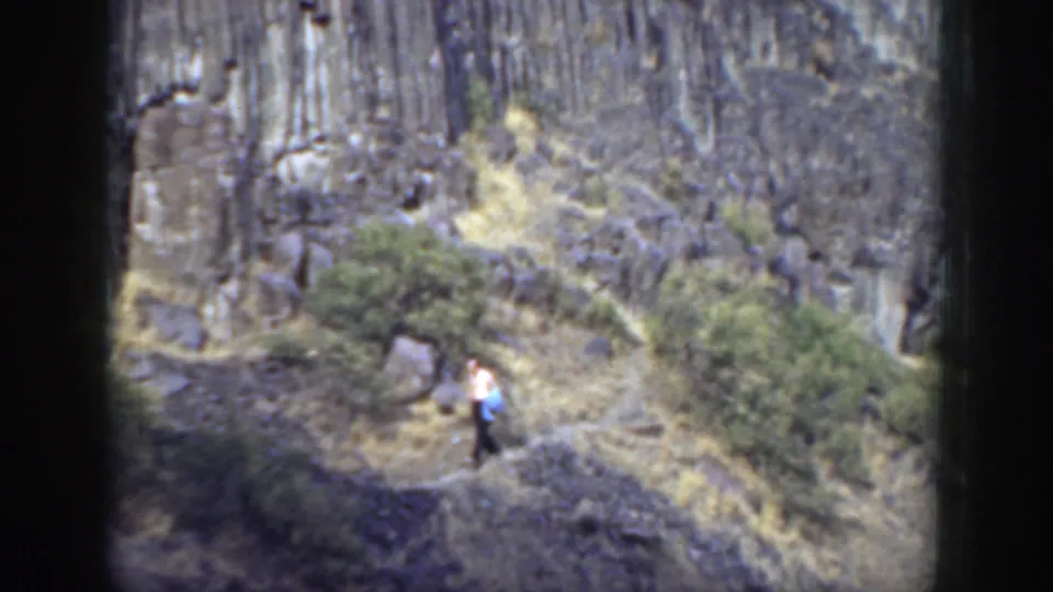 The hiker moves along the California mountain path, carrying a blue bag against a background of shrubs, dry grass, and dark rock.