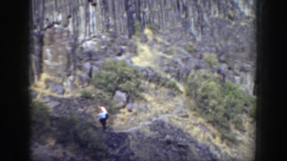 The man appears lower on the trail, walking through dry grass and brush below the rocky cliff in archival 8mm footage.