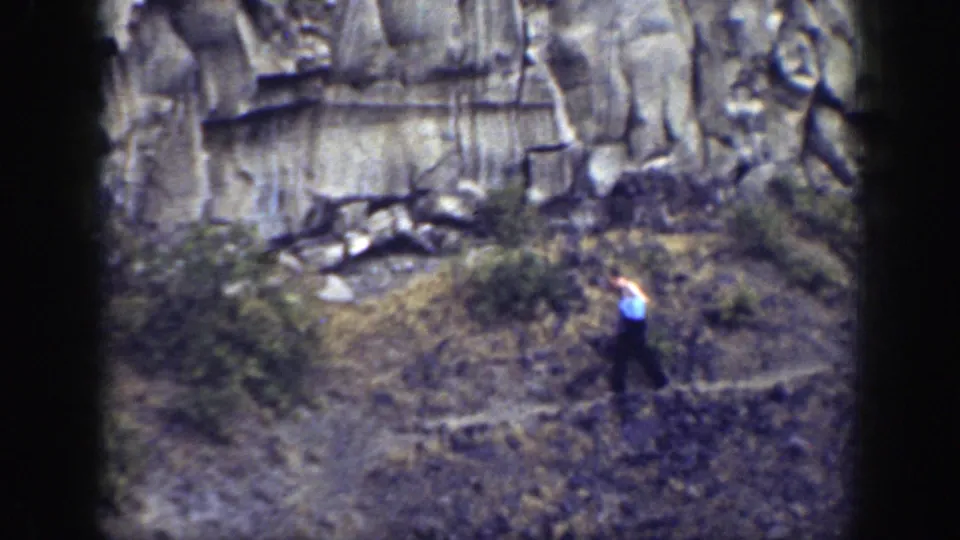 A man in a light shirt and dark trousers carries a blue bag while walking through dry brush below a rugged California cliff face.