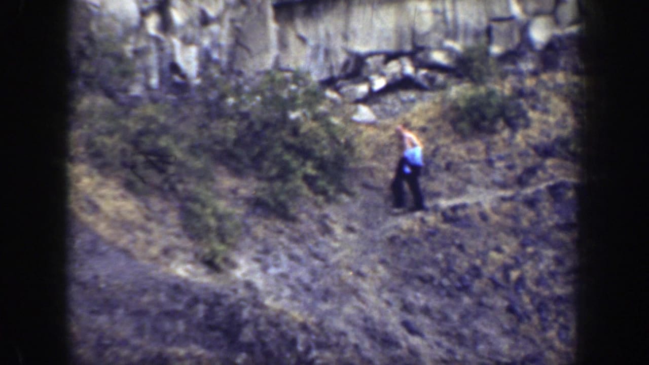 Shirtless man carrying blue cloth walks down a rocky slope below a cliff.