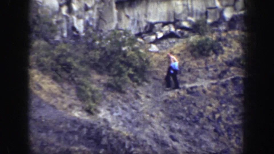 In 1937 California home-movie footage, a shirtless man with a blue cloth moves carefully across a rocky slope beneath a cliff face.