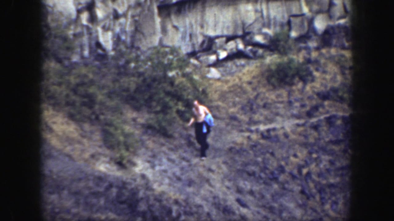 Man stands on uneven rocky ground with brush and cliff rock behind him.