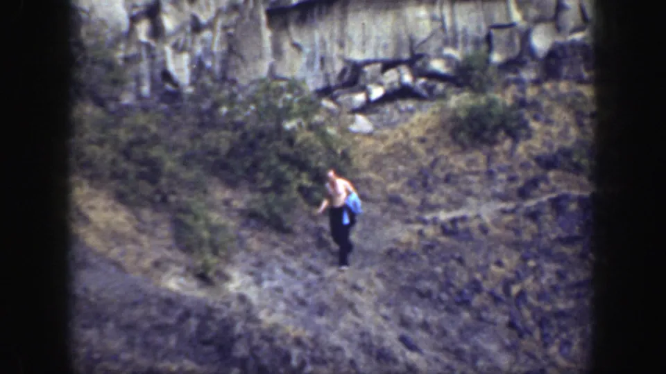 The man continues along the dry rocky hillside, holding a blue cloth with brush and a pale cliff wall behind him.