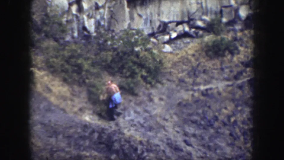 The shirtless man has moved farther down the California hillside, seen near the edge of the rocky grade with brush above him.