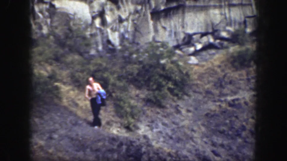 Near the end of the clip, the man appears lower on the path, carrying the blue cloth below shrubs and exposed cliff rock.