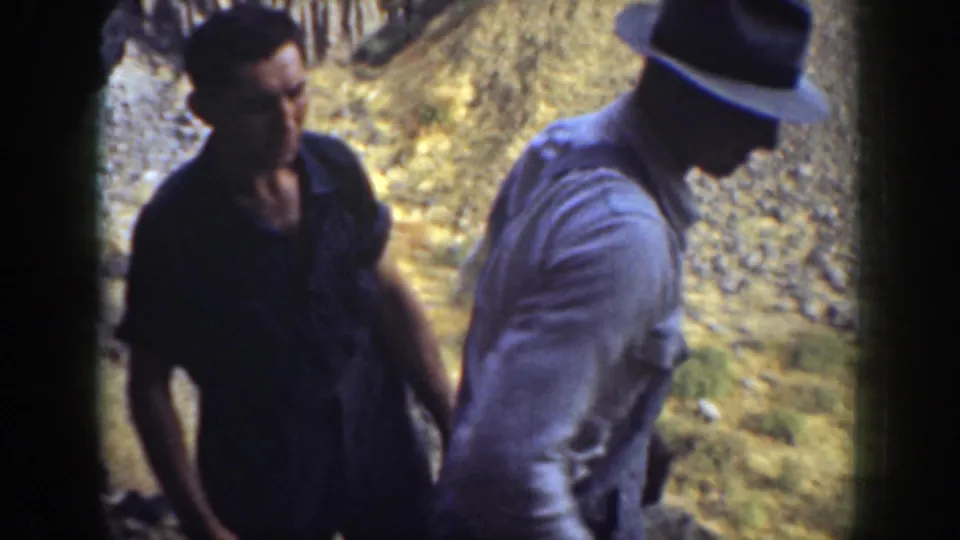 1937 California home-movie frame with two men in casual shirts, one wearing a hat, on a dry rocky slope.