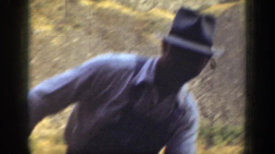 Close handheld 8mm frame of a man in a brimmed hat and light shirt near a sunlit California rock face.