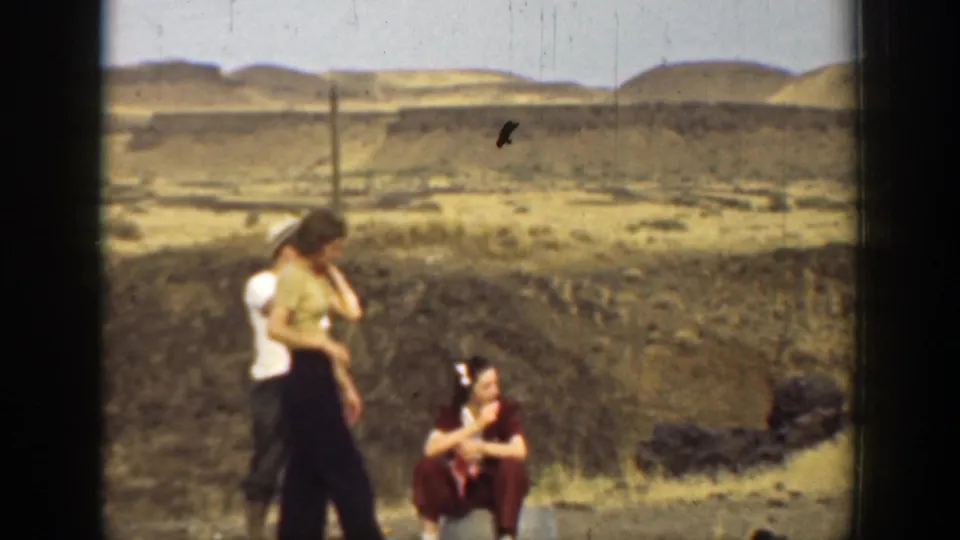 1937 California home-movie frame of companions outdoors, including two standing figures and a seated woman in a maroon outfit.