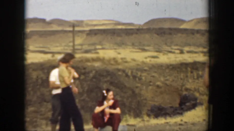 1937 California 8mm frame showing adults talking in a dry outdoor area, with a seated woman, scrubby ground, and distant hills.