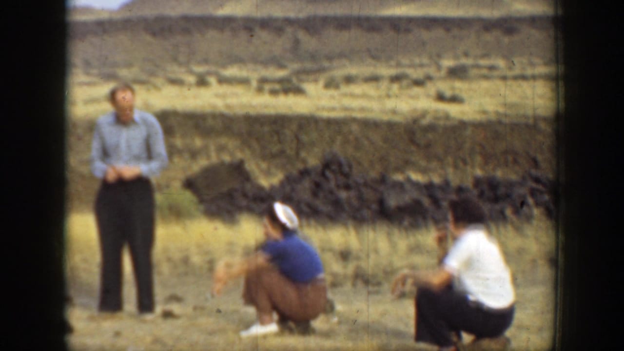 Three adults in a dry landscape, with one standing and two crouching near dark rocks.