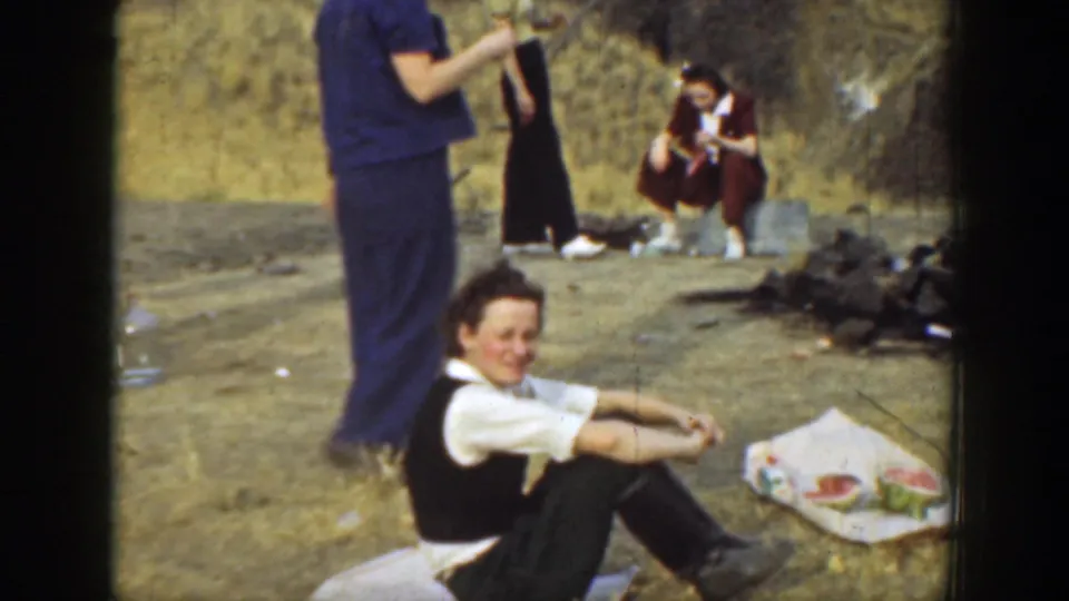 1937 California home-movie frame with a seated adult smiling at the camera during a casual outdoor gathering.
