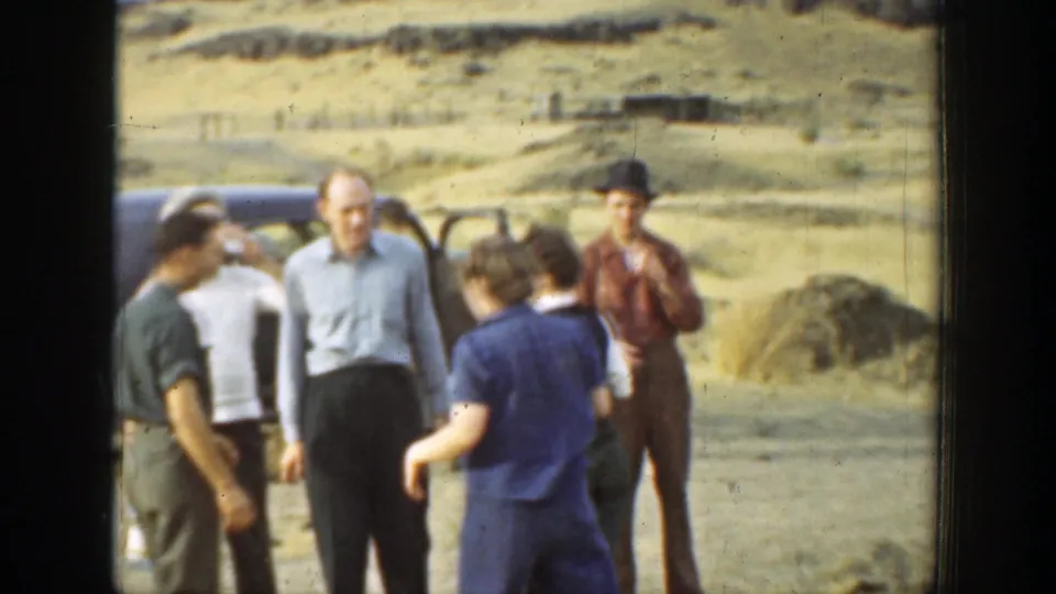 Adults gather near parked cars in a 1937 California hillside setting, with one man facing the camera and others turned in conversation.