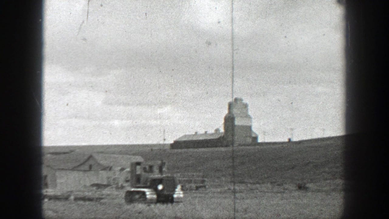 Small tracked tractor in an open field with farm buildings and a tall structure in the distance.
