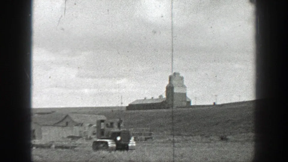 1937 California 8mm frame showing a small tracked tractor crossing open rural land near distant farm buildings and a tall grain-elevator-like structure.