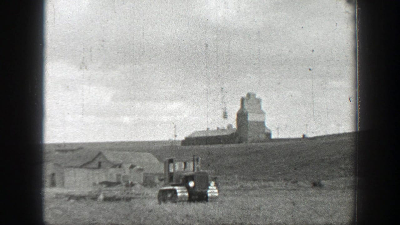 1937 California 8mm frame with a tracked tractor moving across a rural field below distant farm buildings and a tall agricultural structure.
